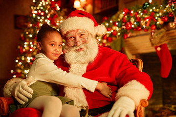 Fototapeta na wymiar Portrait of smiling traditional Santa Claus with cute little girl sitting in his lap by Christmas tree