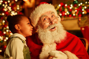 Fototapeta na wymiar Portrait of cute black girl whispering secrets to Santa Claus on Christmas eve