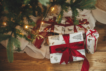Fototapeta na wymiar Stylish christmas gifts under christmas tree with golden lights bokeh. Wrapped christmas presents with red ribbon under decorated tree in room. Atmospheric eve. Holiday banner.