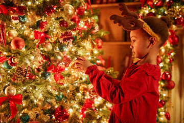 Side view portrait of cute little girl decorating Christmas tree with ornaments and twinkling...