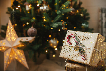 Stylish christmas gifts at christmas tree with golden lights. Wrapped christmas presents with candy cane under decorated tree in room. Merry Christmas and Happy Holidays! Atmospheric banner