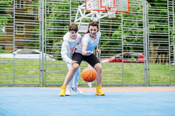 Girl and her younger brother, teenager, play basketball on modern basketball court under open sky