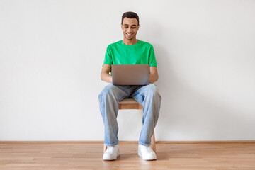 Cheerful arab male freelancer using laptop computer, sitting on chair over white wall and typing on keyboard, copy space