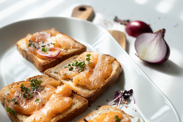 delicious slices of toasted bread with tasty slices of salmon and radish sprouts on them, white plate on a white table in blurred onions in the background, sunny kitchen on a summer day