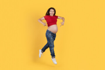 Fototapeta na wymiar Joyful Young Pregnant Woman Pointing At Her Belly While Jumping In Air