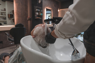 Detalle de peluquero lavando el pelo a un cliente senior, en la peluquería. - obrazy, fototapety, plakaty