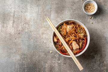 Funchose salad with chicken and vegetables in bowl with wooden chopsticks on green background, top view with text space. Asian food menu
