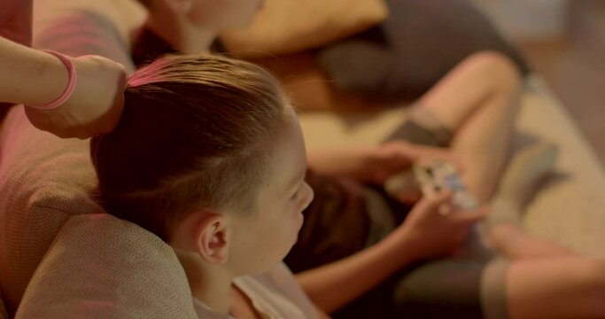 Fixing hair on the head of a teenager. Hands pull a bundle of hair, holding an elastic band. Whiskey and the back of the head are shaved. In the background, a boy plays a controller in bokeh.