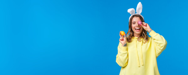 Holidays, traditions and celebration concept. Happy cheerful young pretty female celebrating Easter day, showing two painted eggs and laughing, wearing cute rabbit ears, blue background