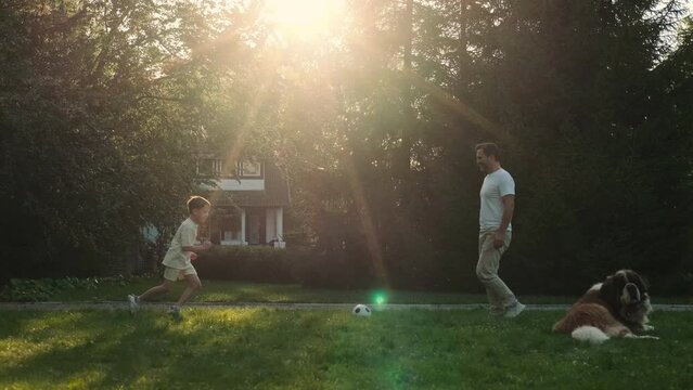 Dad and son play soccer ball on the lawn during sunset.