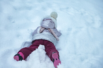 Fototapeta na wymiar Little girl having fun lying on snow in winter public park in warm wear, rear view. Cute child in bright winter clothes walking in city snowy garden. Concept snow wintertime and childhood. Copy space