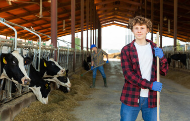 Portrait of confident smiling teen boy working in outdoor cowshed, standing with tool near stall...