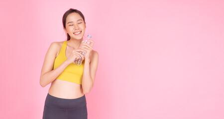 Sporty asian woman holding fresh pure water in bottle after workout standing on pink background. Slim fit body young girl wear sportswear drink fresh water for healthy over isolated pastel background.
