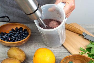 woman prepares a healthy detox drink in a blender - a green smoothie with fresh fruits, blueberries, green spinach and kiwi.