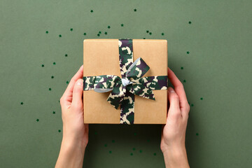 Female hands holding gift box with camouflage military ribbon bow over green background, Defender of the Fatherland Day, USA Veterans Day holidays celebration.