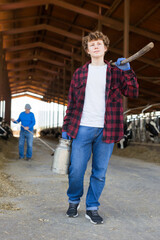 Young and old men working in cowhouse. Young man carrying milk can and working tool in hands, older one cleaning floor of cowshed in background.