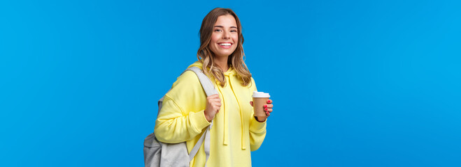 Joyful young blond female student with backpack smiling camera satisfied as holding cup of...