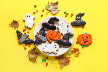 Plate with Halloween cookies, fallen leaves and candies on yellow background