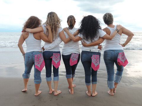 women hugging back to back in denim jeans and pink bandanas on the beach, pinktober