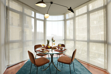 Beautiful circular beveled edge clear glass dining table with crockery of fresh fruit and sweet desserts, brown velvet upholstered chairs and bay window with blinds