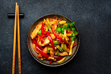 Asian cuisine, stir fry with chicken, red paprika pepper and zucchini bowl. Black kitchen table background, top view, copy space