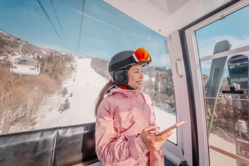 Foto op Aluminium Skivakantie - Vrouwenskiër die telefoonapp in gondelskilift gebruikt. Meisje glimlachend met behulp van mobiele smartphone met skikleding, helm en bril. Ski winter activiteit vakantie concept. © Maridav