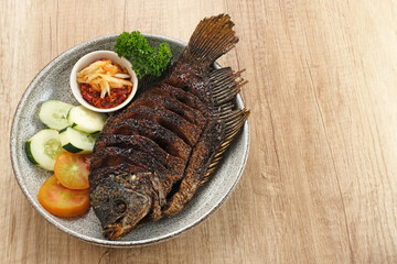 Gurame Bakar Madu, Grilled Gourami with honey and soy sauce. Served with sambal (chilli sauce), cucumber and tomatoes in white plate.   

