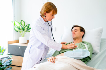 Senior doctor use stethoscope to check body health of young man patient who lie on bed and look like also have problem with arm stay in patient room.