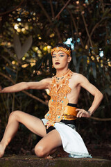 Asian man sitting down on his knee while wearing a gold costume and golden crown on the top of the...