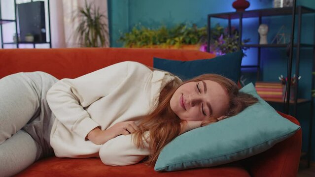 Tired caucasian adult girl lying down in bed taking a rest at home. Carefree young woman napping, falling asleep on comfortable orange sofa with pillows. Closed her eyes enjoy daytime nap alone