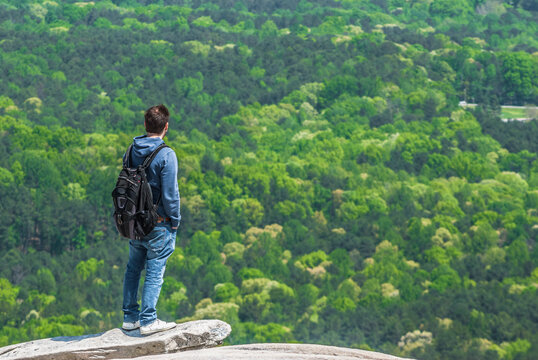A young man with a backpack standing on the edge of the cliff and looking at the green forest below