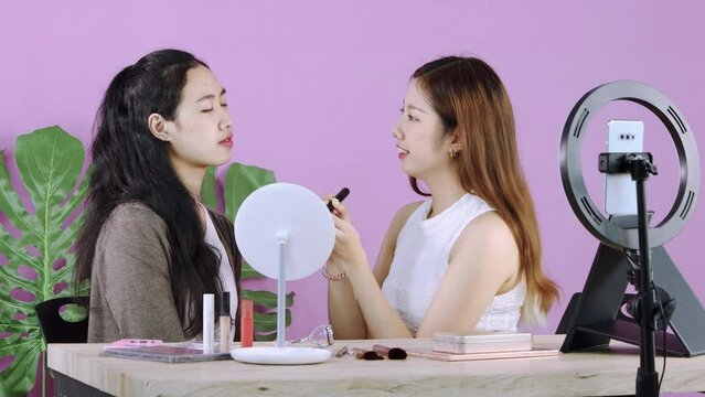 Young asian makeup artist woman live streaming how to make up with pretty woman infront of smartphone cemera while recording her blog on pink color background. 4K video.