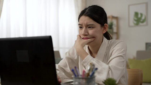 frowning Chinese businesswoman working from home is holding face and looking at laptop screen while thinking about difficult decision at work in the living room
