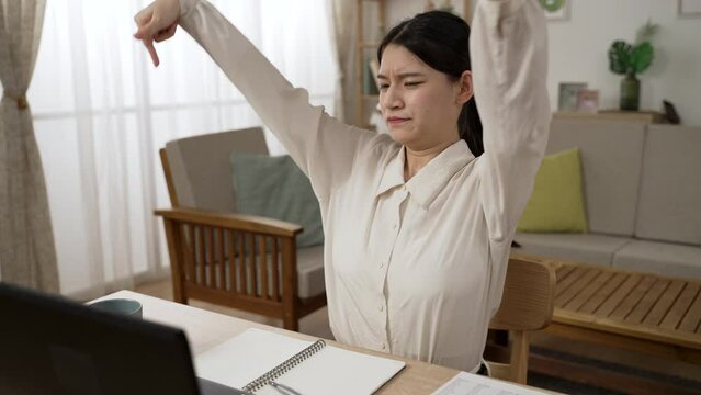 stressed Asian female working from home is stretching her stiff body muscles and sitting back in chair looking into distance with arms behind head to relax