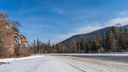 The highway goes around a cliff with steep slopes. Trees grow in snowdrifts on the roadsides. A mountain range against the blue sky. Altai. Chuysky tract