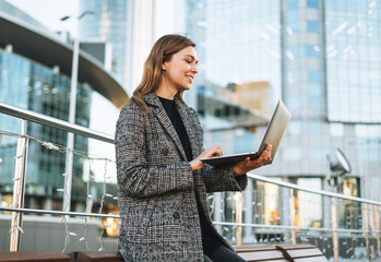 Young smiling woman in coat using laptop in evening city street