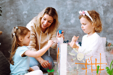 Cute educator help little girls make artistic craft in playroom of kindergarten. Kids sitting and...