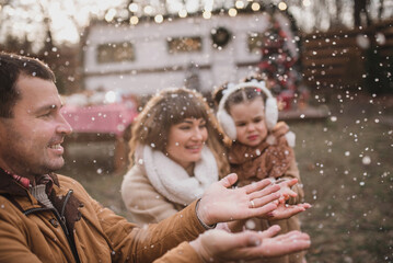 Obraz premium Happy family with two daughters dressed in stylish fur coats a play hugging and enjoying near the trailer in the forest with New Year's decorations. Selected focus