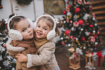 two little twin girls dressed in stylish fur coats play, hug and enjoy near the trailer in the forest with New Year's decorations. Family decorating the Christmas tree