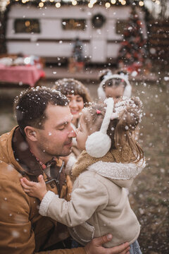 Happy family with two daughters dressed in stylish fur coats a play hugging and enjoying near the trailer in the forest with New Year's decorations. Selected focus