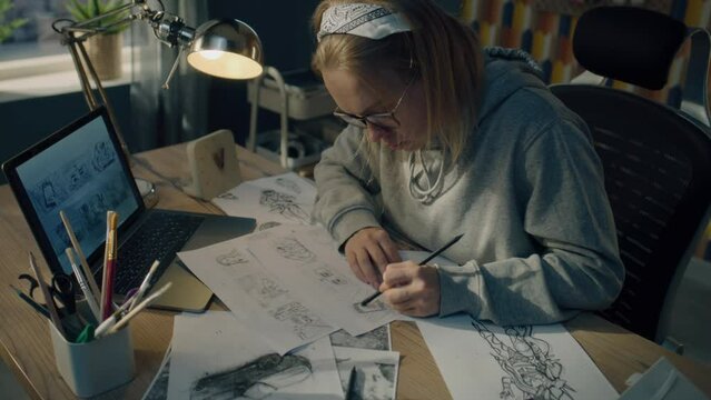 Female animator creates sketches for the cartoon. Looks at the laptop screen infront of her and draws on the paper. Comics Compilation. Storyboard movie layout for pre-production.