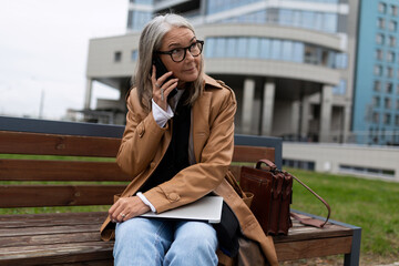 Business elderly businesswoman talking on a mobile phone with a laptop on her lap against the backdrop of a business center