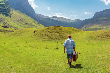 Fototapeta na wymiar A man with a suitcase walks through grass in the mountains. Interesting journey of a lonely men