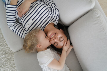 Adopted girl child kiss smiling mother, hugging on cozy sofa at home. Happy motherhood, adoption