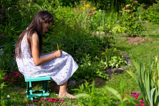 Young caucasian girl in a white dress is drawing on a plein air sitting on a folding chair in summer garden 