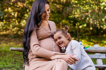 Fototapeta premium Happy pregnant mother and daughter hugging on a park bench. Happy motherhood. Beautiful woman with a little daughter in nature