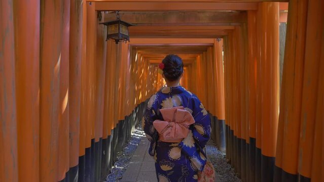 Cinematic shot of a Japanese woman dressed in a traditional Japanese cloth walking through a way surrounded by thousands of red Toris (Japanese spiritual gate). Few tourists are in the distance.