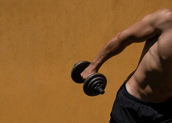 Young man working out lifting dumbbell outdoors during midday with harsh sunlight and yellow...