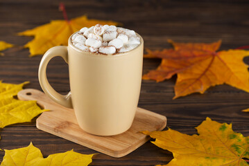 Hot chocolate with marshmallows in a mug on a wooden table against the background of autumn leaves.