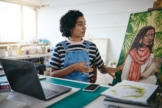 Art girl and laptop video call in studio for professional remote sale of painting on canvas online. Creative indian woman with focus and concentration selling artwork on the internet in workshop.